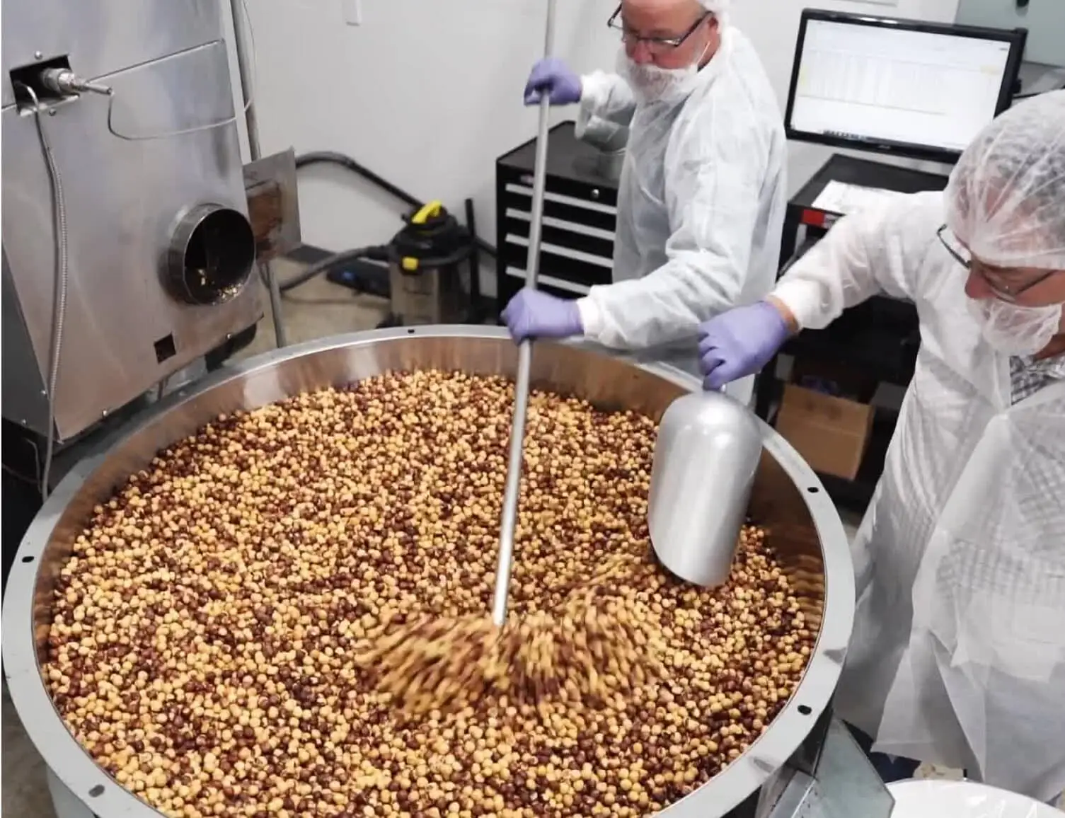 Roasted nuts are being mixed in a large metal bowl by workers in a food processing facility.