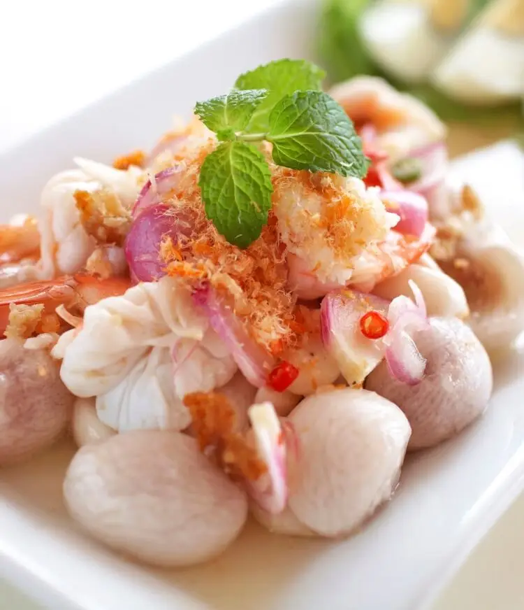 Seafood salad rests on a white plate. Shrimp and onions are visible. A sprig of mint decorates the top.